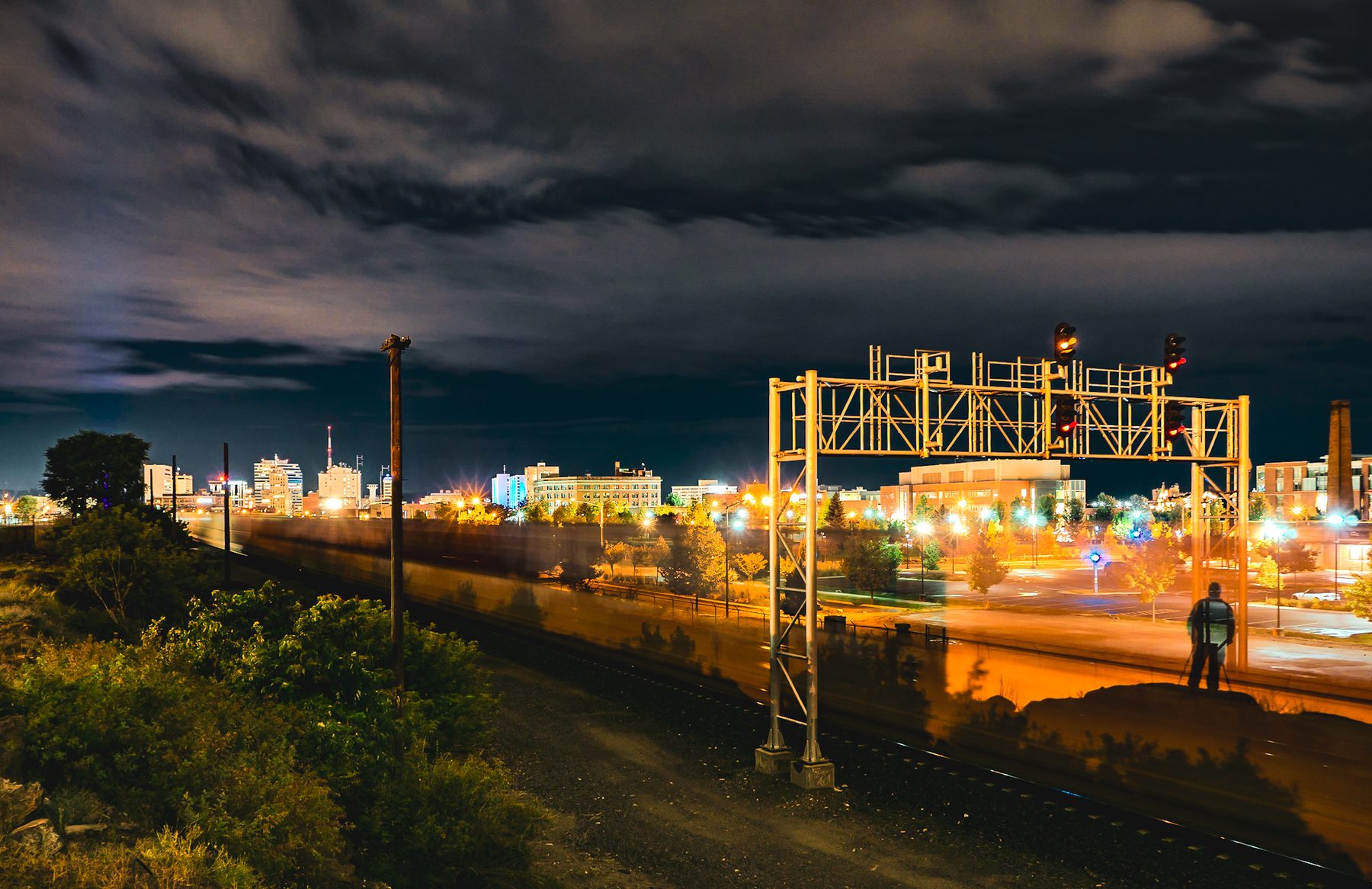 Long-exposure night photograph: a signal bridge, city skyline, a train blurring past, and a lone photographer at a tripod trackside